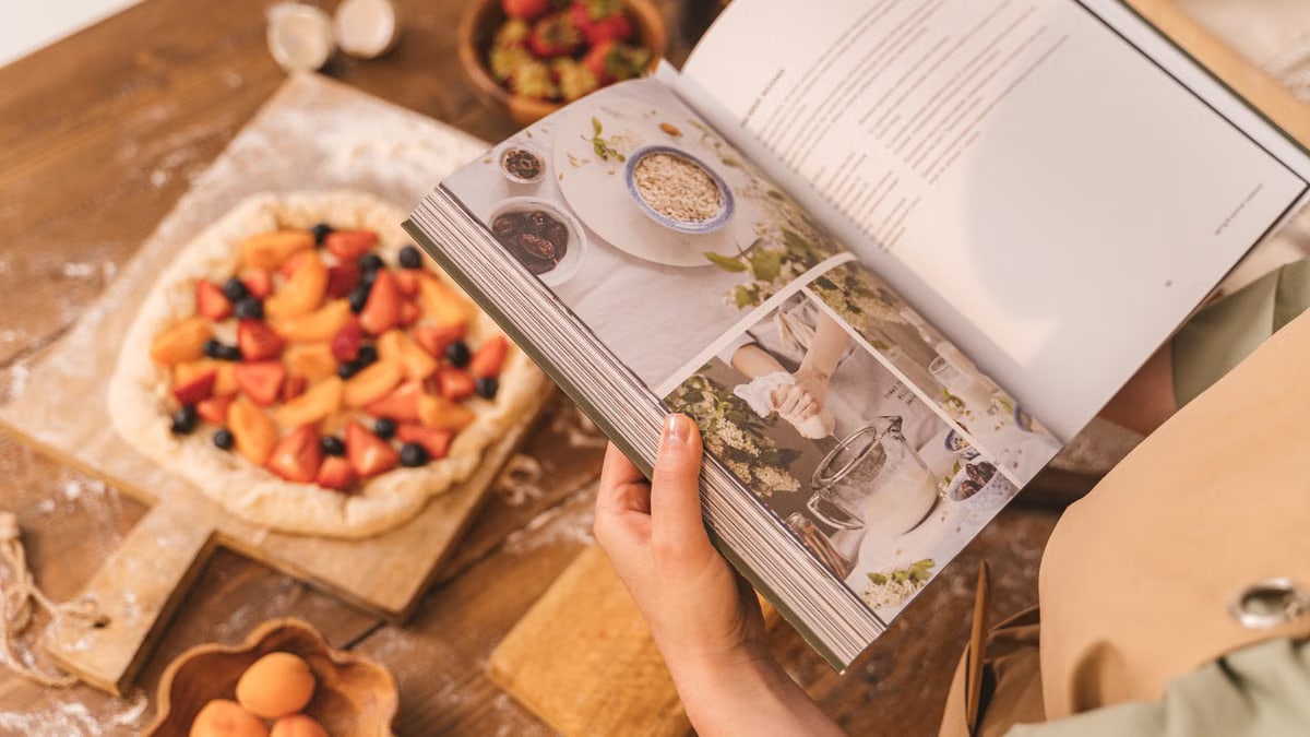 A person reads a cookbook while preparing a fruit tart on a wooden table.